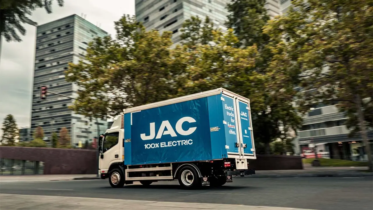 Cami&oacute;n de transporte el&eacute;ctrico de JAC en una ciudad moderna con edificios y &aacute;rboles.